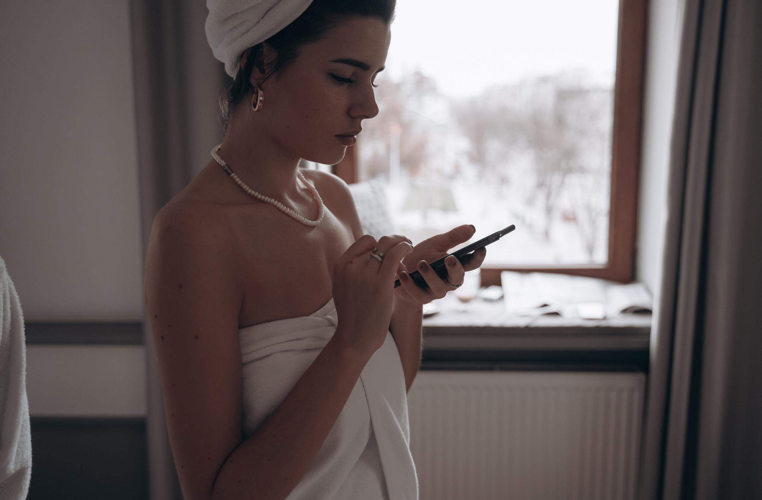 Žena sa peškirom oko glave i tela koristi mobilni telefon, pretražujući sadržaj o nezi kože.